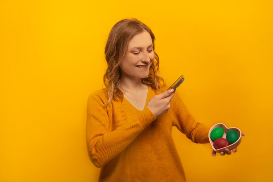 Happy smiling girl in yellow sweater holding heart shaped plate saucer with three multicolored painted easter eggs, taking photo of dyed easter eggs isolated on yellow background.

Easter Day concept.