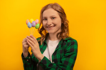 Happy smiling girl holding small multicolored painted dyed easter eggs sticks, wearing green checkered plaid shirt with white shirt and celebrating Easter Day isolated on yellow background.