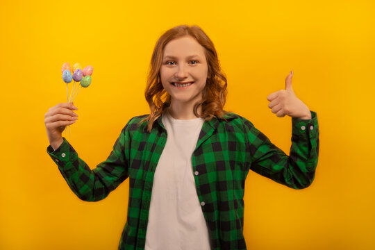 Happy smiling girl holds small multicolored painted dyed easter eggs sticks, shows thumb up or keeps like hand gesture and wears green checkered plaid shirt isolated on yellow background.

Easter Day.