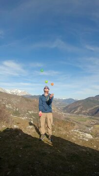 Man juggling 4 balls (yellow, orange, pink, green) at the top of a mountain. He wear sport clothes and sunglasses. Mountain background hilighted. Vertical video in real time ready for slow motion.
