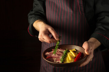 The cook puts rosemary on a plate with sliced steak and cheese. The concept of serving dishes to order by a waiter with a place for advertising on a black background