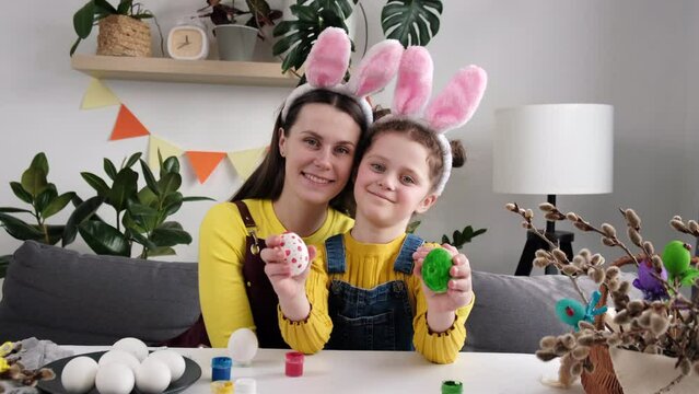 Sweet family portrait of happy young mother and cute little daughter in fluffy bunny ears holding small painted multi-colored Easter eggs, tenderly embracing and smiling in cozy light room at home