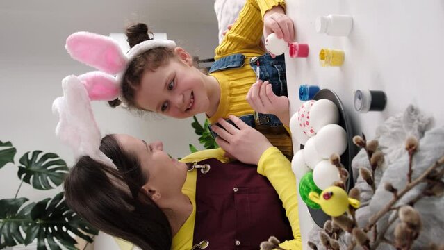 Vertical video of loving young mother teaching pretty daughter painting eggs sit together at table, wears fluffy bunny ears. Easter celebration and traditions. Concept creativity, children's education