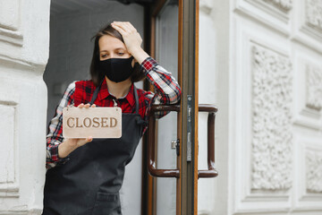 sad woman with closed sign in her hands at restaurant or bar wearing protective mask. working hours...
