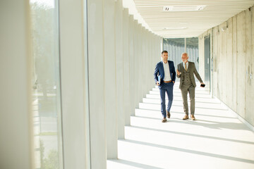 Young and a senior businessman walk down an office hallway, deep in conversation