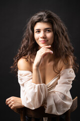 Naklejka premium Classic studio portrait of a young brunette in a white loose top, who is sitting on a chair against a black background.