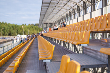Rows of yellow chairs in a stadium in perspective with people in the background.