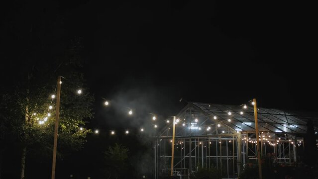 A garland of lamps illuminates the yard. Outdoor patio lights on gloomy evening, a small greenhouse in the background, slow motion shot. Wedding decoration.