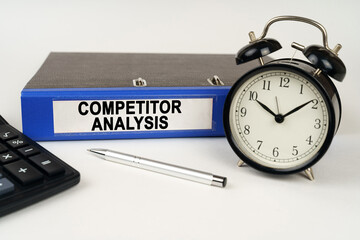 On a white surface, an alarm clock calculator and a folder with the inscription - COMPETITOR ANALYSIS