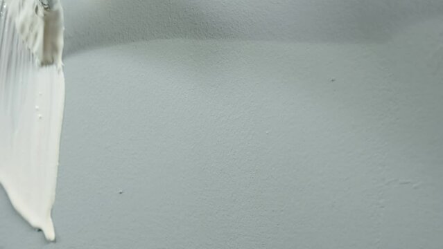 The unrecognizable man paints a gray wall with white paint using a yellow brush. Close up of a hand of painter or repairman sliding a brush over a wall and painting over it. The concept of repair.