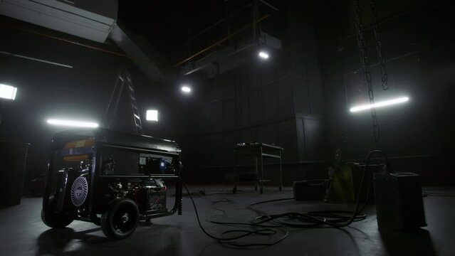 Black mobile gasoline power generator in a dark workshop. Gasoline powered generator that produces light. Battery equipment connected by wires. Dark empty room with light.
