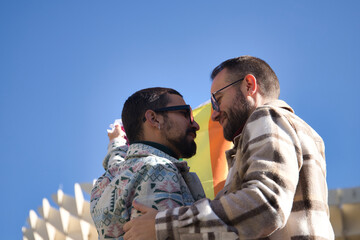 Real marriage of gay couple, looking at each other while holding a gay pride flag, happy and complicit. Concept lgtb, lgtbiq+, couples, in love, pride, flag.