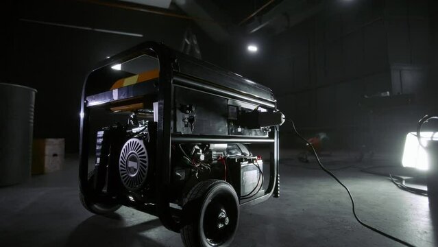 Black mobile gasoline power generator in a dark workshop. Gasoline powered generator that produces light. Battery equipment connected by wires. Dark empty room with light.