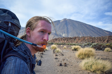 Pretty young traveler man hiking drinking water on mountain with Teide volcano on the background. Enjoying hot weather and sun in Spain. Staying hydrated on sport hike. Hydration bladder.