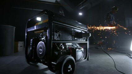 Black mobile gasoline power generator in a dark workshop. In the background, a man in blue overalls, a hard hat and goggles cuts metal with a grinder or a circular knife. In the process, sparks fly.