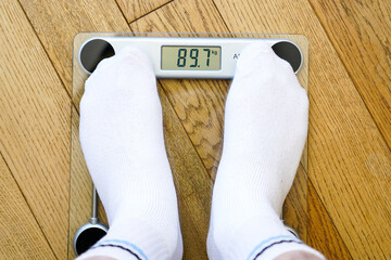 A man in white socks stands on a transparent electronic floor scale, the weight reading is 89 kg