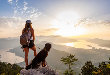 A sporty girl with a backpack stands on the edge of a mountain with a Rottweiler dog