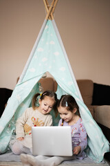 Two girls sisters watching on laptop at wigwam tent. Technology and home concept. © AS Photo Family