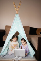 Two girls sisters watching on laptop at wigwam tent. Technology and home concept. © AS Photo Family