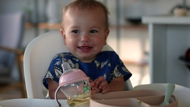 Happy baby girl 1 year old looks towards camera in high chair and giggles - close up on face