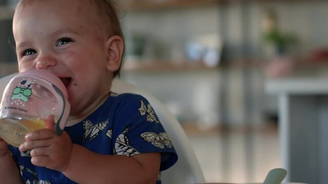 Baby girl drinking from sippy cup in highchair smiles and laughs towards camera - close up on face