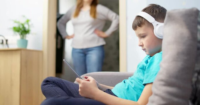 Mother scolds her son. Boy uses tablet with headphones and ignores his mom