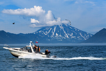 Fototapeta premium A boat with tourists floats in the Pacific Ocean against the backdrop of a snow-covered volcano and mountains. Kamchatka.