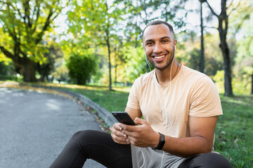 A young African-American male sportsman, athlete, runner in headphones sits in the park on the curb. Holds the phone in his hands, listens to music, calling, rest. He looks at the camera, smiles.