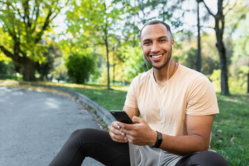 Fototapeta premium A young African-American male sportsman, athlete, runner in headphones sits in the park on the curb. Holds the phone in his hands, listens to music, calling, rest. He looks at the camera, smiles.