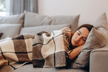 Beautiful serene woman sleeping on comfy sofa under warm cozy plaid in modern living room. Healthy daytime nap, tiredness relief, repose and relaxation during day, lazy weekend at home.