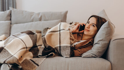Lazy weekend. The girl is lying on the sofa under a blanket and talking on the phone.