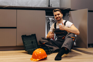 A young plumber holds a wrench in his hand and smiles at the camera on the background of the kitchen.