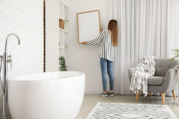 Young woman hanging blank frame on white brick wall in bathroom