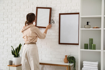 Young woman with hammer hanging blank frame on white brick wall in bathroom