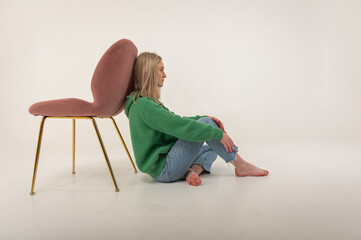 portrait of a young emotional girl sitting on the floor near a chair in a flirtatious mood, gesturing with her hands, empty space, wearing jeans and a green sweater n, space for text