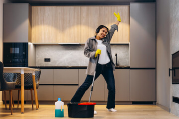 Happy woman cleaning home, singing at mop like at microphone and having fun, copy space. Housework,...