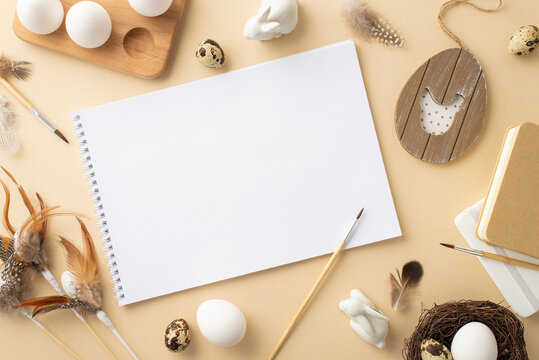 Easter concept. Top view photo of sketchbook paintbrushes quail eggs wooden egg holder rustic decor nest ceramic bunnies and quail feathers on isolated beige background with blank space - Powered by Adobe