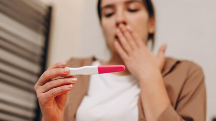 Young worried sad girl sitting in the bathroom and holding a positive pregnancy test in her hands. She is in shock, this is an unexpected pregnancy. The concept of different pregnancy emotions.