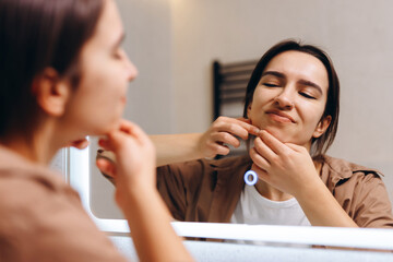 A young girl stands in front of a mirror in the bathroom and presses a pimple on herself chin.
