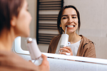 A young girl stands in front of a mirror in the bathroom and rinses her mouth with an irrigator.
