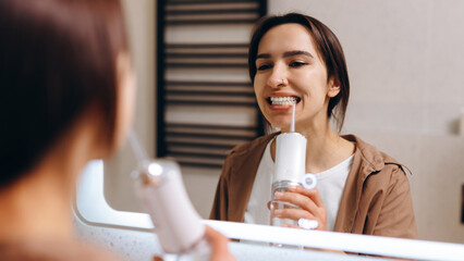 A young girl stands in front of a mirror in the bathroom and rinses her mouth with an irrigator.