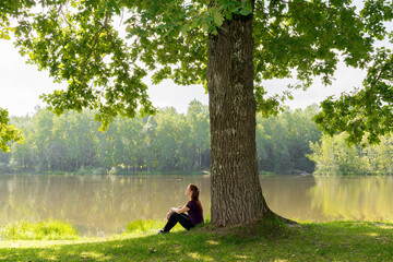 Teen young girl woman sitting on grass under huge high big old oak,trunk tree near lake,river, meditating,relaxing and smiling in National Park.inspiration nature beauty harmony landscape outdoors
