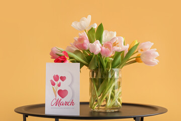 Vase with tulips and greeting card for Women's Day on table near yellow wall