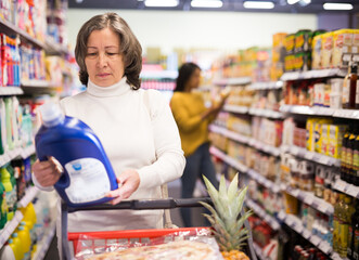 Portrait of focused elderly woman shopping in supermarket, reading product label while choosing detergents in household chemicals department