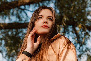 Caucasian young pretty stylish woman standing street pier near big huge tree dressed brown trench coat hold handbag smiling poses outside city, spring autumn season. Cute brunette hair lady
