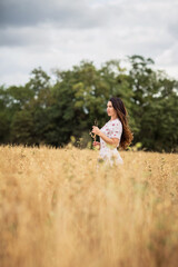 Naklejka premium A beautiful brunette woman in a white dress runs along a field of golden chickpeas ear. Stylish girl in the field in windy weather