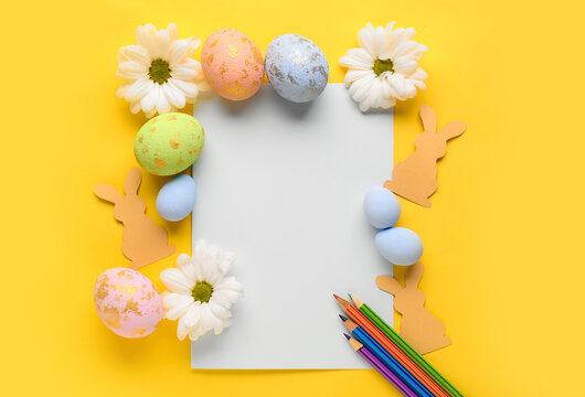Blank paper sheet with pencils, Easter eggs, rabbits and daisy flowers on yellow background