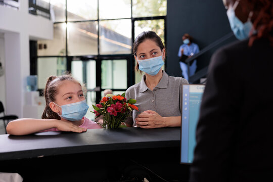 Reception Worker Talking With Mother And Kid Explaining Medical Expertise During Checkup Visit Consultation In Hospital Lobby. People Wearing Protective Face Mask To Prevent Infection With Covid19