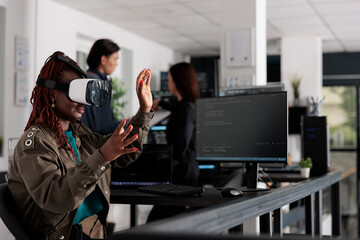African american system engineer wearing virtual reality goggles working to develop security program in it big data office, software developer with vr headset coding html script with database.