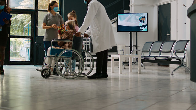 Young Kid Visiting Old Woman In Wheelchair During Covid 19 Pandemic, Bringing Flowers To Elderly Patient With Disability. Mother And Child Coming To See Person With Physical Impairment.